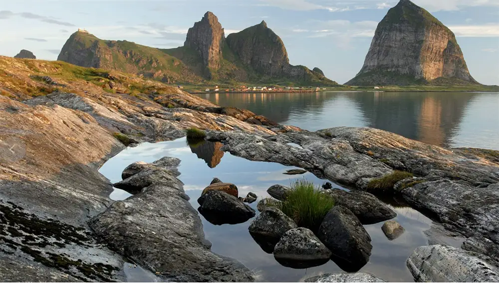 A breathtaking view of the Archipelago on Norway’s remote Helgeland coast. Towering granite peaks rise dramatically from the calm, reflective waters, creating an otherworldly landscape. In the foreground, weathered rock formations and tidal pools mirror the sky, while a small village nestled at the base of the mountains highlights the isolation and raw beauty of this Arctic island paradise
