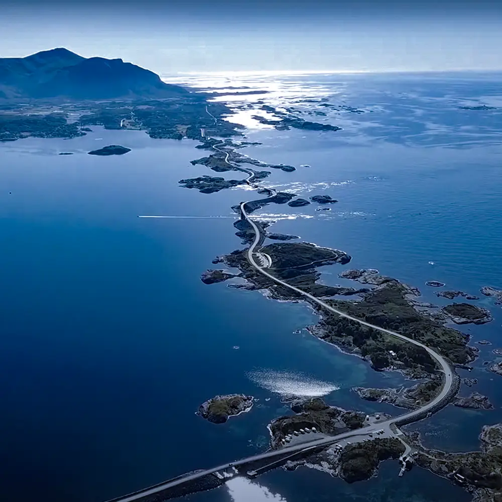 A stunning aerial view of Norway’s Atlantic Road, an iconic coastal highway winding across small islands and skerries, connected by sweeping bridges. The road appears to float above the deep blue sea, with the sun casting a shimmering glow over the vast ocean and distant mountains.