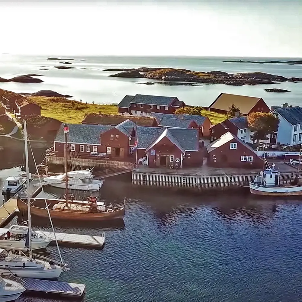 A serene coastal scene of Håholmen, a historic fishing village in Norway, bathed in warm evening light. Sailboats and traditional wooden buildings line the waterfront, with small islands and calm waters stretching into the distance. The soft glow highlights the rugged beauty of this remote Nordic outpost.