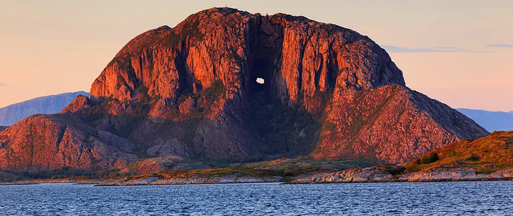 A breathtaking view of Torghatten, the legendary mountain in Norway known for its massive, naturally-formed hole piercing through its center. Bathed in the warm glow of the setting sun, the rugged rock face contrasts with the deep blue waters below, creating an awe-inspiring landscap