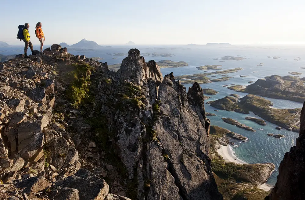 Two hikers stand atop the rugged peak of Rødøyløva, gazing out over Norway’s Helgeland coast. Below them, a maze of tiny islands and turquoise waters stretches to the horizon, bathed in the golden light of the Midnight Sun. The steep cliffs and dramatic terrain make this one of the most rewarding hikes in the region, offering an unparalleled view of the Arctic landscape.