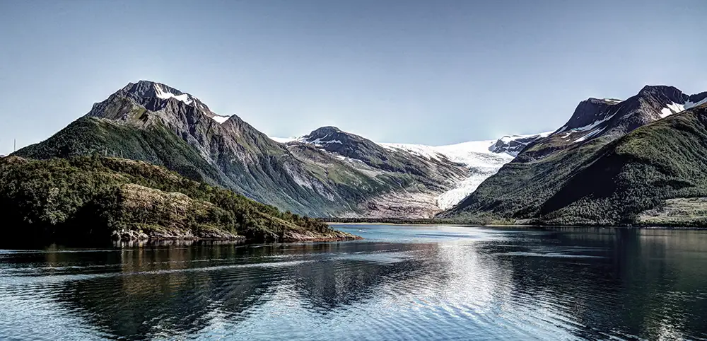 A striking view of Svartisen Glacier, Norway’s second-largest glacier, cascading down between rugged mountain peaks into a glassy fjord. The deep blue water reflects the surrounding landscape, creating a scene of pure Arctic beauty. Lush greenery in the foreground contrasts with the icy expanse, highlighting the dramatic meeting of land, sea, and ice in this remote and untouched region.