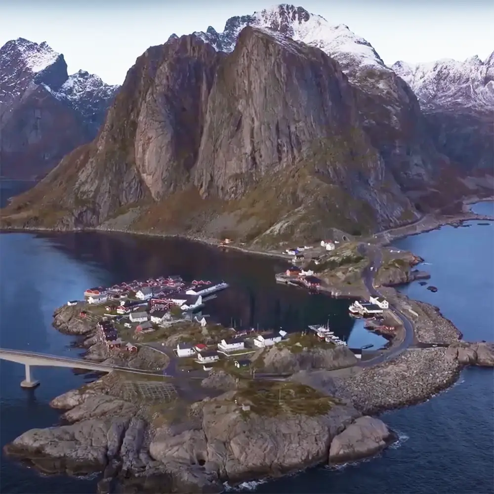 A stunning aerial view of a remote Norwegian fishing village nestled at the foot of towering, snow-capped peaks. The dramatic landscape features rugged cliffs, deep fjords, and traditional red and white houses scattered along the shoreline. A bridge connects the island to the mainland, showcasing the region's breathtaking blend of nature and human resilience.