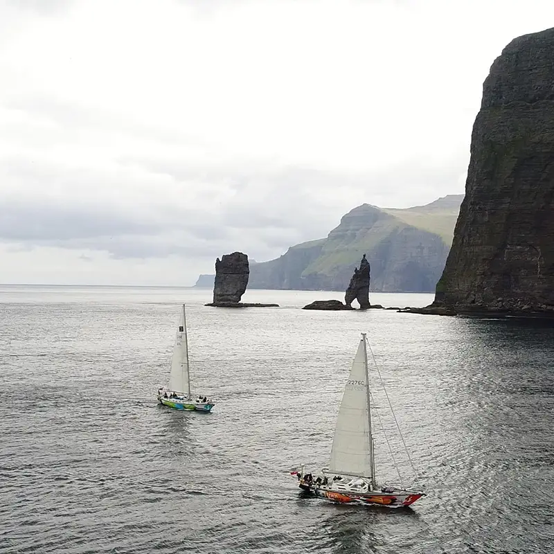 Two sailboats navigate the waters of the Faroe Islands, passing towering cliffs and dramatic sea stacks rising from the ocean. The rugged coastline in the background is shrouded in mist, adding to the remote and wild atmosphere. The boats, their sails full of wind, contrast against the dark waters, embodying the spirit of adventure sailing in one of the world’s most breathtaking maritime landscapes.
