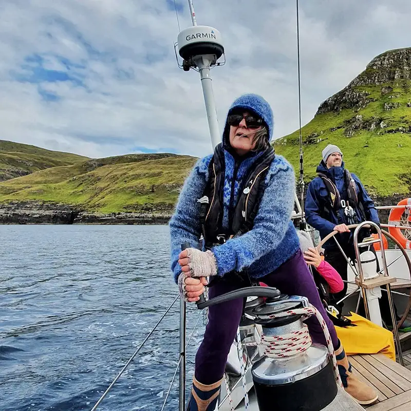 A woman in a blue wool sweater, gloves, and sunglasses operates a winch on a sailboat in the Faroe Islands, surrounded by dramatic green cliffs. Dressed for the cool North Atlantic weather, she looks focused as she trims the sails. Behind her, other crew members, also in warm gear, assist with sailing, enjoying the rugged, remote scenery. The deep blue water contrasts with the lush, rolling hills, creating a striking backdrop for this adventure sailing experience.