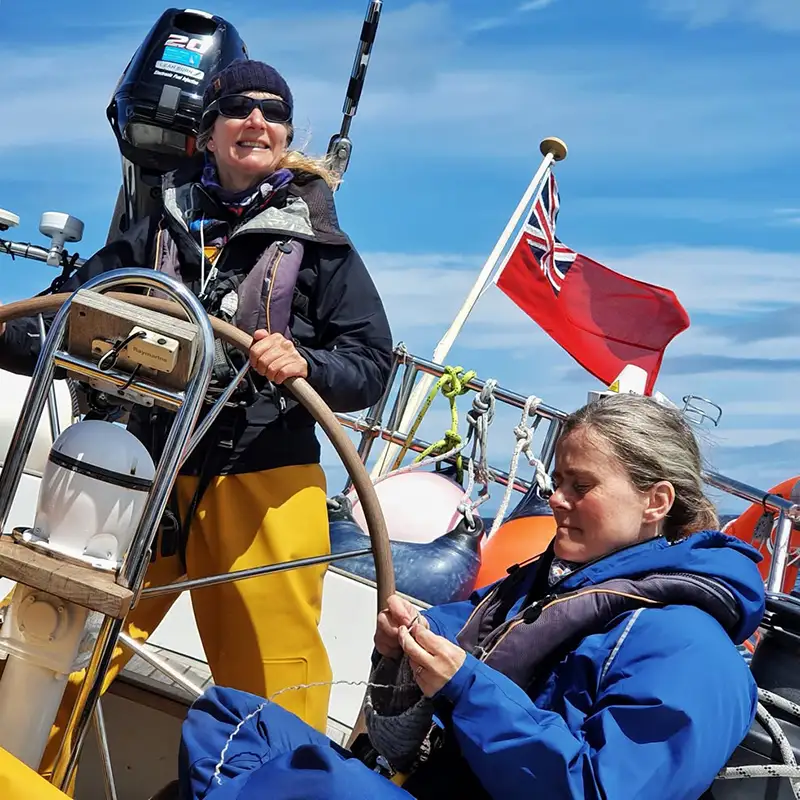 Two women sailing in the Faroe Islands, one at the helm steering the boat with a smile, while the other focuses on a task with rope. Dressed in warm sailing gear and life jackets, they navigate under clear blue skies with a Red Ensign flag flying at the stern. The scene captures the essence of adventure sailing, teamwork, and the joy of exploring the North Atlantic Ocean by yacht.