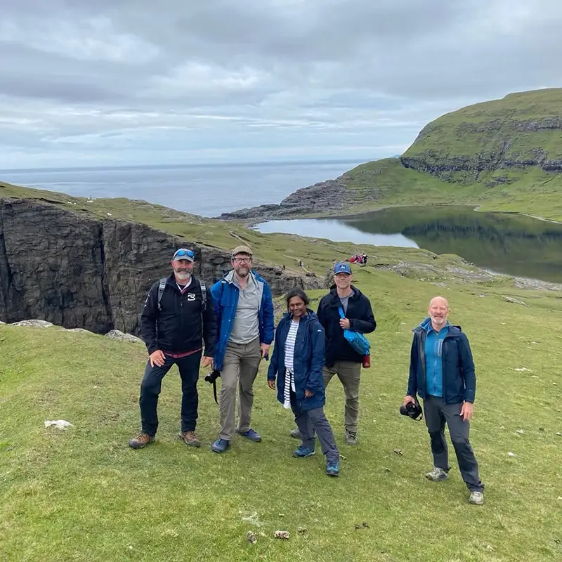 A group of hikers stands on a grassy hillside in the Faroe Islands, smiling after a trek through the rugged landscape. Behind them, a dramatic cliff drops off into the North Atlantic Ocean, while a serene lake reflects the overcast sky. The surrounding green slopes and distant coastline capture the wild beauty of the Faroese wilderness, making this a perfect stop for adventure seekers exploring the islands by land and sea.
