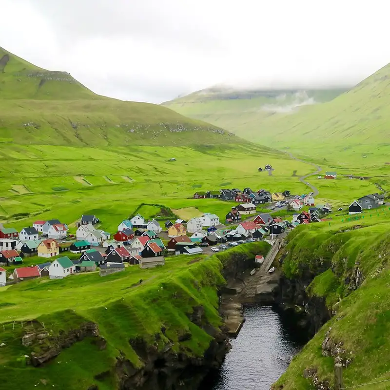 The charming village of Gjógv in the Faroe Islands, nestled between rolling green hills and a dramatic natural harbor. Colorful houses with traditional Nordic architecture dot the landscape, contrasting against the lush grass and rugged cliffs. A narrow inlet cuts through the land, leading out to the North Atlantic Ocean, while mist lingers over the distant peaks, adding to the remote and picturesque atmosphere.